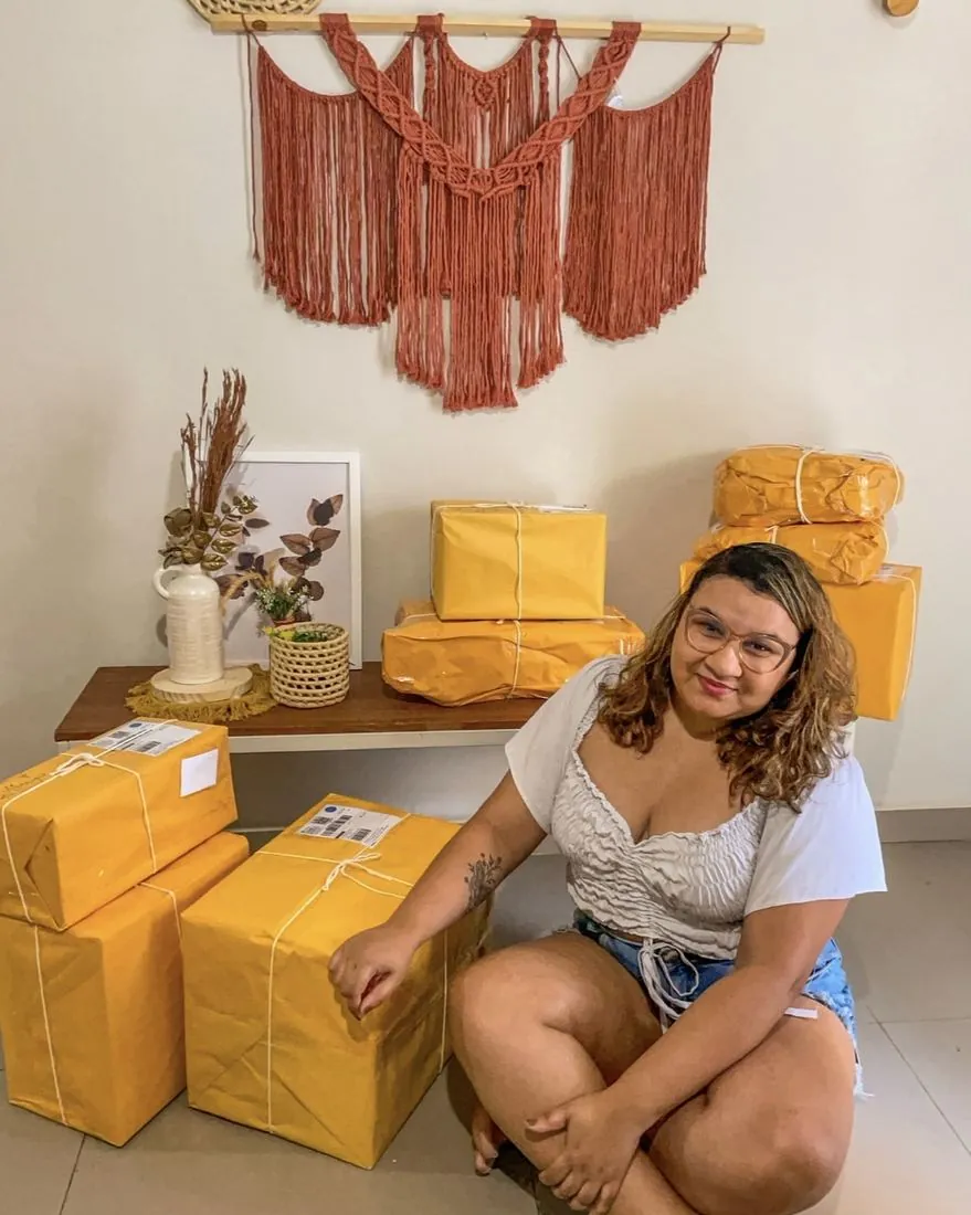 Lari, da Alarí Handmade, sentada entre caixas de encomendas com painel de macramê ao fundo